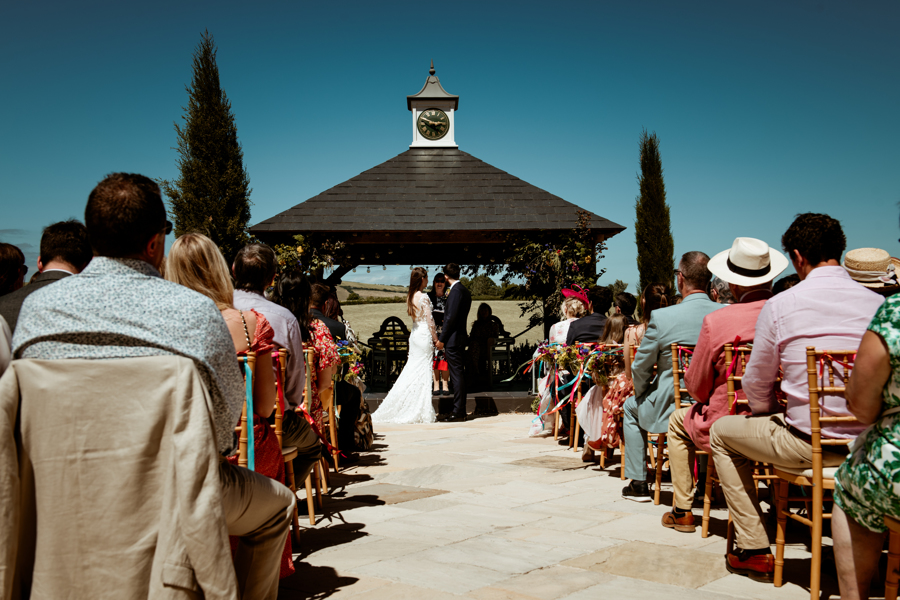 Fran and James standing in front of the Clock House on the terrace at Harefield Barn exchanging their vows with fields and a beautiful blue sky beyond and guests watching on.